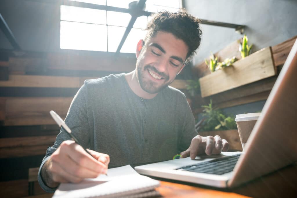 Gelukkige ondernemer bij zijn klein bedrijfsrestaurant die op een laptop en een blocnote werkt die zeer gelukkig glimlachen terwijl het genieten van een koffie